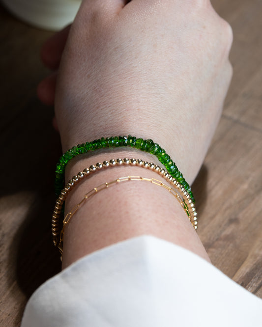 Bright green chrome diopside beaded gemstone bracelet stacked on a wrist with 14k gold-filled paperclip chain, and 14k gold-filled beaded bracelet.