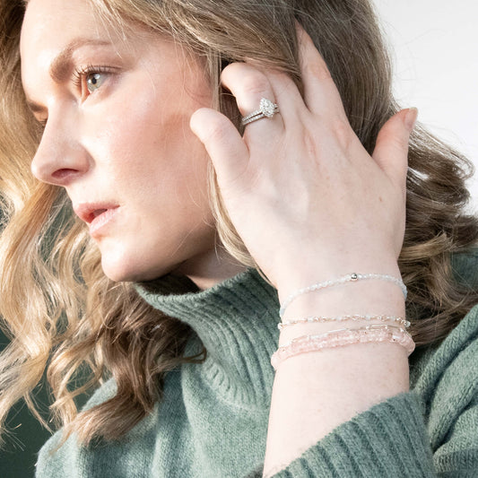 A close-up lifestyle shot of a woman wearing Morganite silver bracelets and and a silver ring with a clear gemstone, styled with a sage green knit turtleneck sweater.