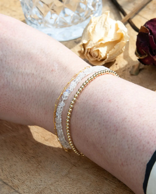 Clear white Moonstone faceted beaded bracelet with flashes of blue stacked on wrist with with 14k gold-filled beaded bracelet and 14k gold-filled paperclip chain on wood table