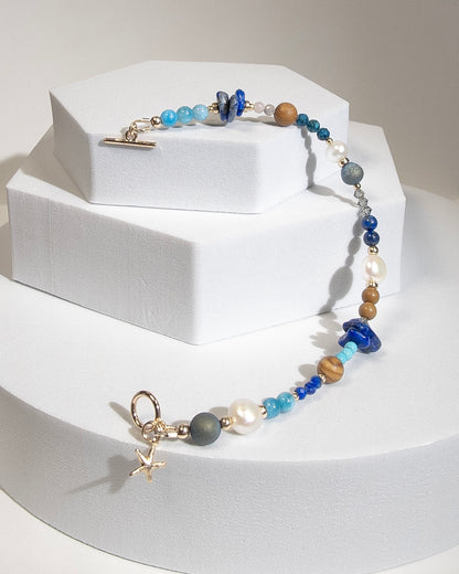 A beaded bracelet with assorted shapes and sizes of Lapis Lazuli, Button Pearl, Wood Jasper, Sunstone, Turquoise, Druzy Quartz, Apetite, and Preciosa Crystal, separated by 14k gold-filled accents beads. Finished with a 14k gold-filled toggle clasp and starfish charm on a geometric white platform