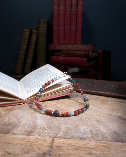 A beaded gemstone bracelet with assorted shapes and sizes of red, green, and brown Jasper, deep purple-pink Portuguese Agate, pale green aventurine, and orange Spessartine Garnet, separated by sterling silver beads. Finished with a sterling silver spring ring clasp resting on an open book on a wood table