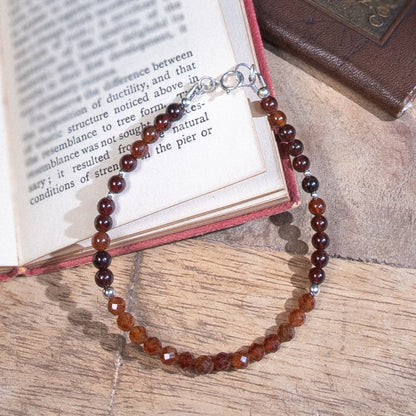 A Spessartine Garnet beaded bracelet with deep orange smooth and faceted round beads with sterling silver accents beads and a spring ring clasp, rested on an open book on a wood table.
