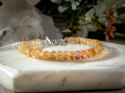 Closeup of orange and yellow Imperial Topaz faceted gemstone beaded bracelet in gradient with sterling silver heart-shaped charm, and toggle clasp on light marble coaster