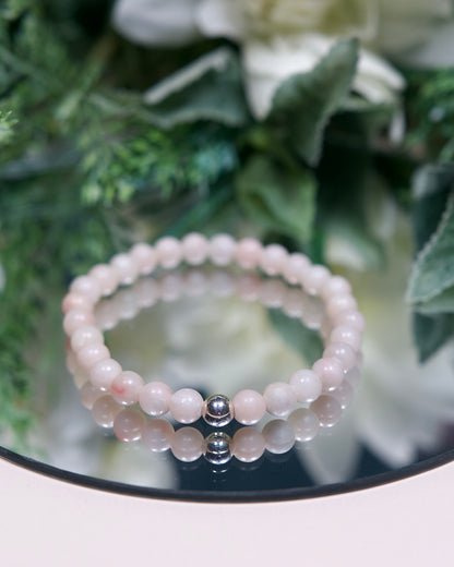 A pale pinkish-white Pink Opal beaded gemstone bracelet with smooth round beads and a sterling silver accent bead on a mirrored background