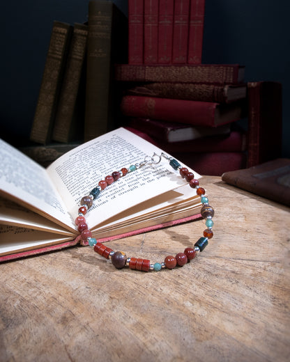 A beaded gemstone bracelet with assorted shapes and sizes of red, green, and brown Jasper, deep purple-pink Portuguese Agate, pale green aventurine, and orange Spessartine Garnet, separated by sterling silver beads. Finished with a sterling silver spring ring clasp resting on an open book on a wood table
