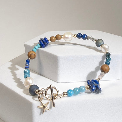 A beaded bracelet with assorted shapes and sizes of Lapis Lazuli, Button Pearl, Wood Jasper, Sunstone, Turquoise, Druzy Quartz, Apetite, and Preciosa Crystal, separated by 14k gold-filled accents beads. Finished with a 14k gold-filled toggle clasp and starfish charm on a geometric white platform