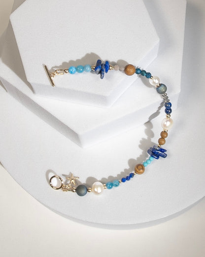 A beaded bracelet with assorted shapes and sizes of Lapis Lazuli, Button Pearl, Wood Jasper, Sunstone, Turquoise, Druzy Quartz, Apetite, and Preciosa Crystal, separated by 14k gold-filled accents beads. Finished with a 14k gold-filled toggle clasp and starfish charm on a geometric white platform