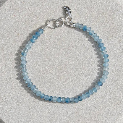 Top down photo of Aquamarine beaded gemstone bracelet with light and dark blue faceted roundelle shaped beads with sterling silver leaf-shaped charm and spring ring clasp on light coaster on a wood table