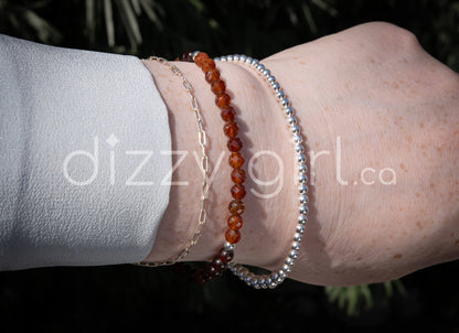 A Spessartine Garnet beaded bracelet with deep orange smooth and faceted round beads with sterling silver accents beads stacked on a wrist with a sterling silver beaded bracelet and a sterling silver paperclip chain