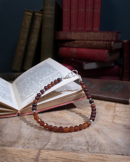 A Spessartine Garnet beaded bracelet with deep orange smooth and faceted round beads with sterling silver accents beads and a spring ring clasp, rested on an open book on a wood table.