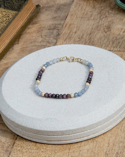 Light blue and dark purple faceted sapphire beaded bracelet with stardust 14k gold-filled accent beads and 14k gold-filled spring ring clasp on light coaster on wood table