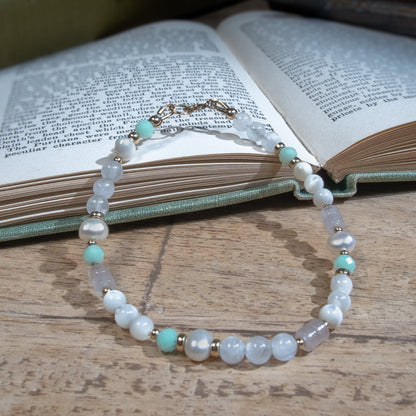 A beaded bracelet with pearlescent white mother of pearl beads, oval-shaped freshwater pearl, opaque white quartz, and pale green jade separated by 14k gold-filled accent beads. Finished with a 14k gold-filled spring ring clasp resting on an open book on a wooden table