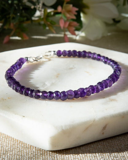A faceted Amethyst beaded gemstone bracelet with a sterling silver spring ring clasp on a white coaster.