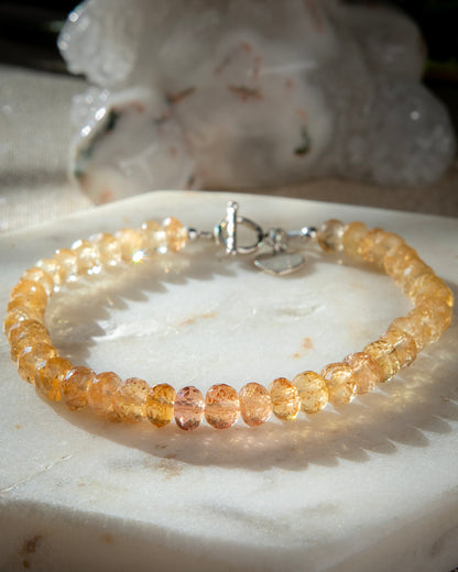 Orange and yellow Imperial Topaz faceted gemstone beaded bracelet in gradient with sterling silver heart-shaped charm, and toggle clasp on light marble coaster