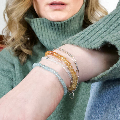 A close-up of a woman wearing a green ribbed turtleneck sweater wearing an Imperial Topaz and other stacked bracelets.