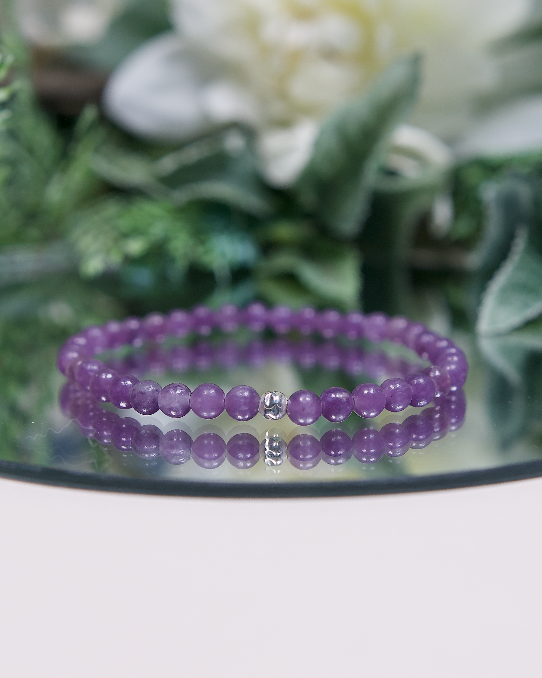 Purple Lepidolite beaded bracelet with a sterling silver accent bead on a reflective surface with green foliage in the background