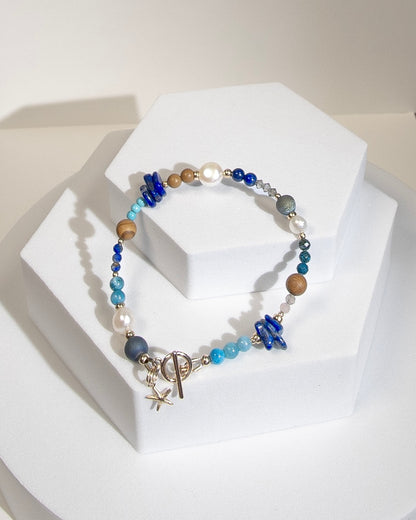 A beaded bracelet with assorted shapes and sizes of Lapis Lazuli, Button Pearl, Wood Jasper, Sunstone, Turquoise, Druzy Quartz, Apetite, and Preciosa Crystal, separated by 14k gold-filled accents beads. Finished with a 14k gold-filled toggle clasp and starfish charm on a geometric white platform
