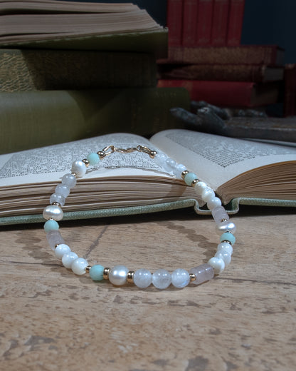 A beaded bracelet with pearlescent white mother of pearl beads, oval-shaped freshwater pearl, opaque white quartz, and pale green jade separated by 14k gold-filled accent beads. Finished with a 14k gold-filled spring ring clasp resting on an open book on a wooden table