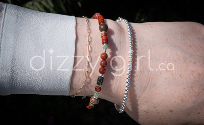 A beaded gemstone bracelet with assorted shapes and sizes of red, green, and brown Jasper, deep purple-pink Portuguese Agate, pale green aventurine, and orange Spessartine Garnet, separated by sterling silver beads, stacked on a wrist with a sterling silver beaded bracelet and sterling silver paperclip chain.