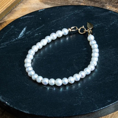 A beaded bracelet made of white freshwater pearls with a gold toggle clasp, displayed on a black surface on a wood table