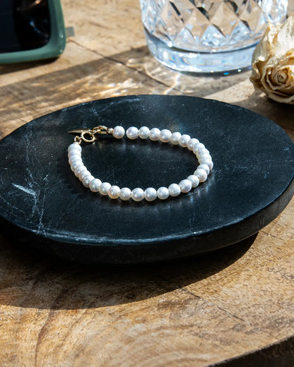 A beaded bracelet made of white freshwater pearls with a gold toggle clasp, displayed on a black surface on a wood table
