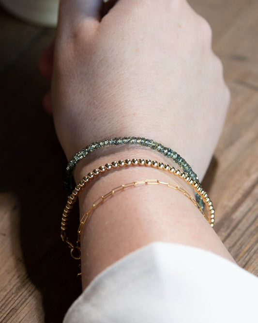 Earthy, muted green Sapphire beaded gemstone bracelet stacked on a wrist with 14k gold-filled paperclip chain, and 14k gold-filled beaded bracelet.