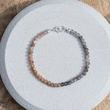 Top down photo of peach and grey moonstone beaded gemstone bracelet with sterling silver spring ring clasp on light coaster on wood table