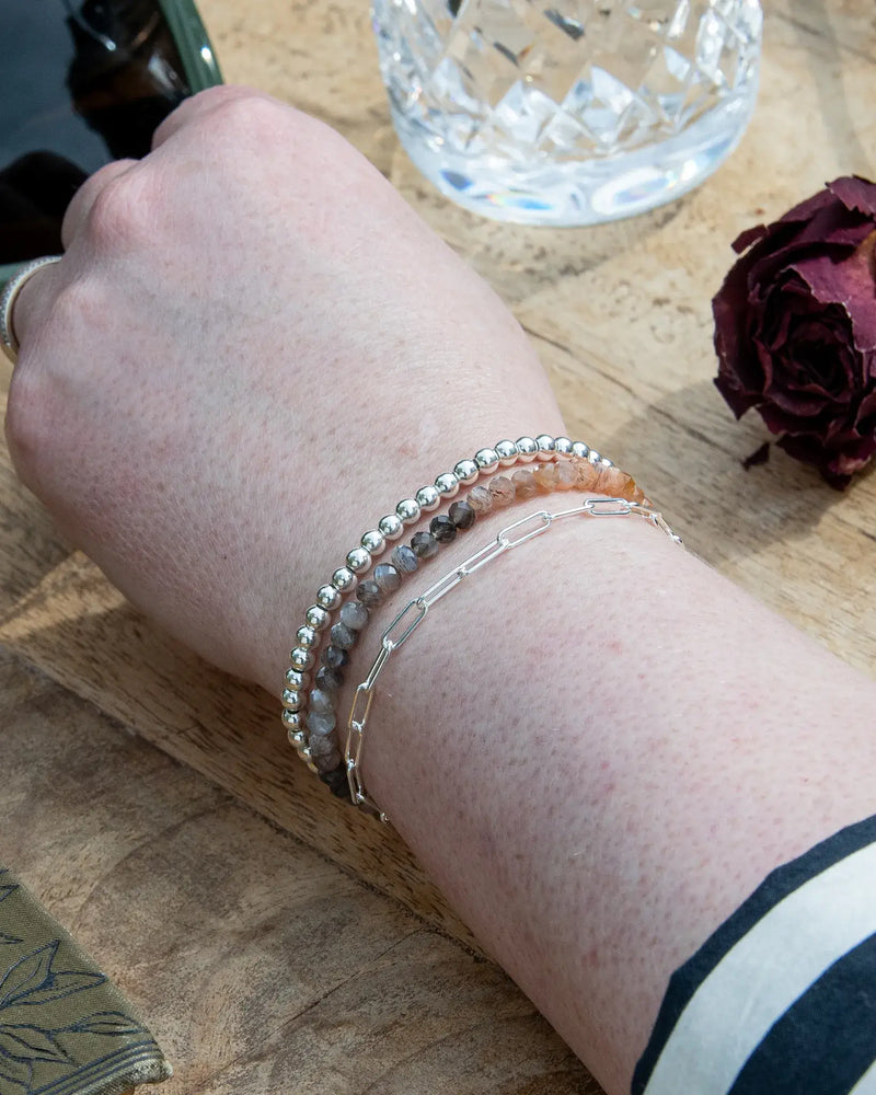 Peach and grey moonstone beaded gemstone bracelet stacked on wrist with sterling silver beaded bracelet and sterling silver paperclip chain on wooden table