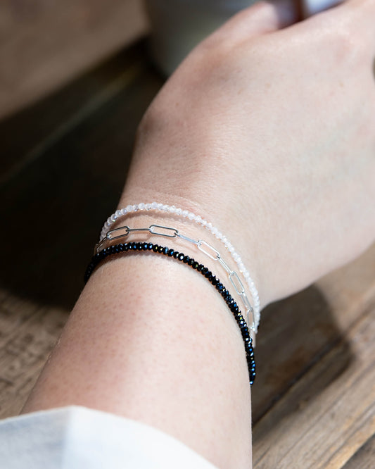 Dainty white zircon beaded gemstone bracelet stacked on a wrist with dark silver hematite and sterling silver paperclip chain bracelets.