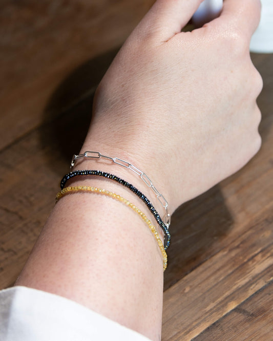 Dainty dark silver hematite beaded gemstone bracelet stacked on a wrist with yellow sapphire and sterling silver paperclip chain bracelets.