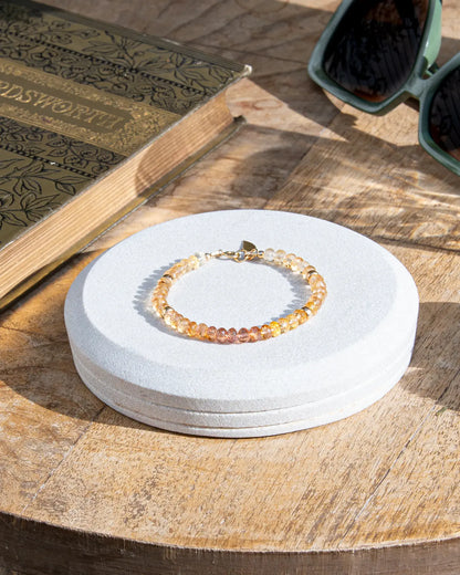 Pink, orange, and yellow Imperial Topaz faceted gemstone beaded bracelet in gradient with 14k gold-filled beads, leaf-shaped charm, and spring ring clasp on light coaster on wood table