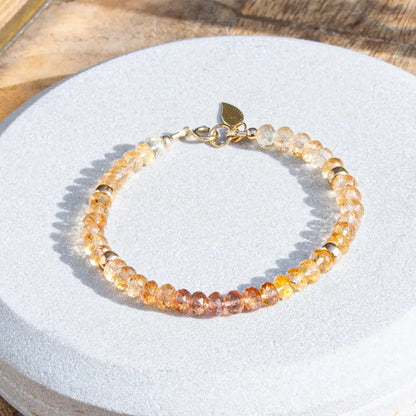 Pink, orange, and yellow Imperial Topaz faceted gemstone beaded bracelet in gradient with 14k gold-filled beads, leaf-shaped charm, and spring ring clasp on light coaster on wood table