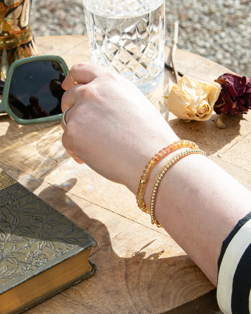 Pink, orange, and yellow Imperial Topaz faceted gemstone beaded bracelet in gradient stacked on wrist with 14k gold-filled beaded bracelet on wood table