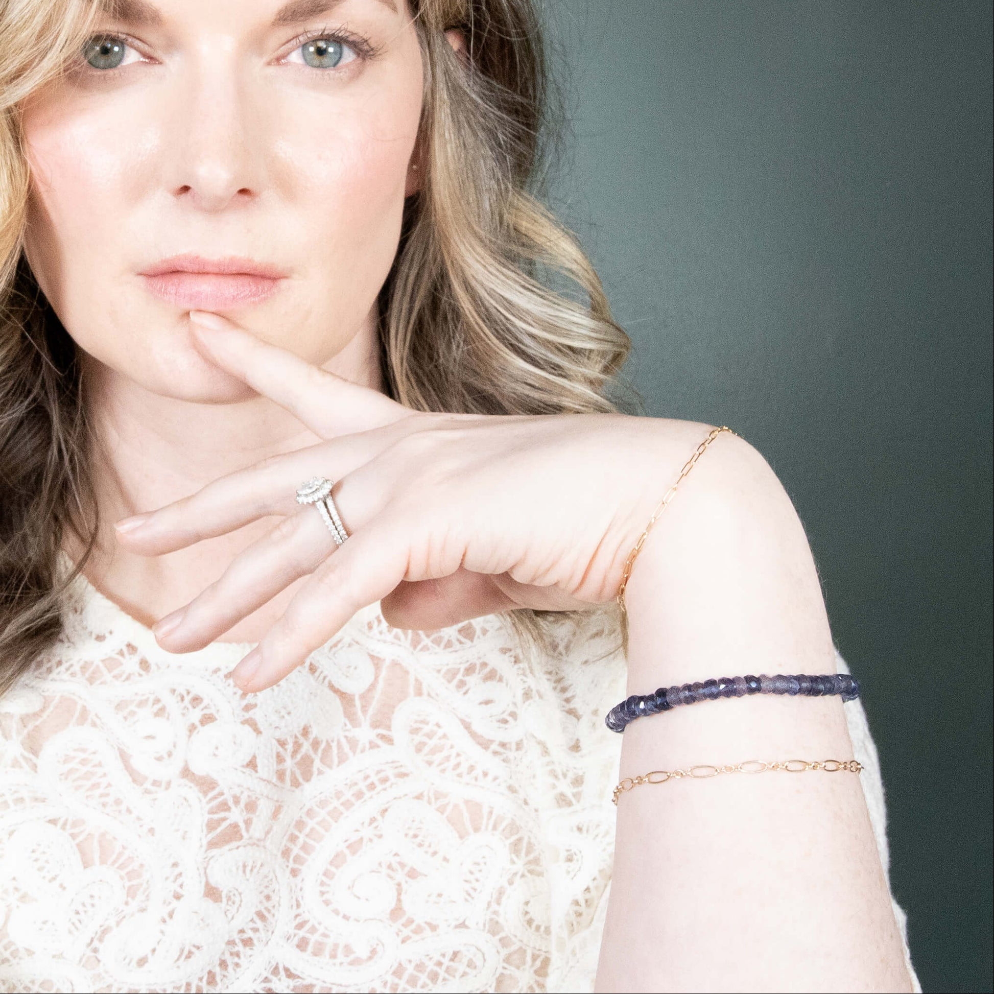A close-up portrait of a woman wearing an Iolite and delicate gold bracelets on her wrist, set against a dark gray background.