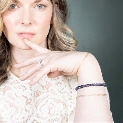 A close-up portrait of a woman wearing an Iolite and delicate gold bracelets on her wrist, set against a dark gray background.