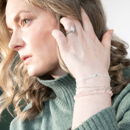A close-up lifestyle shot of a woman wearing Morganite silver  bracelets and and a silver ring with a clear gemstone, styled with a sage green knit turtleneck sweater.