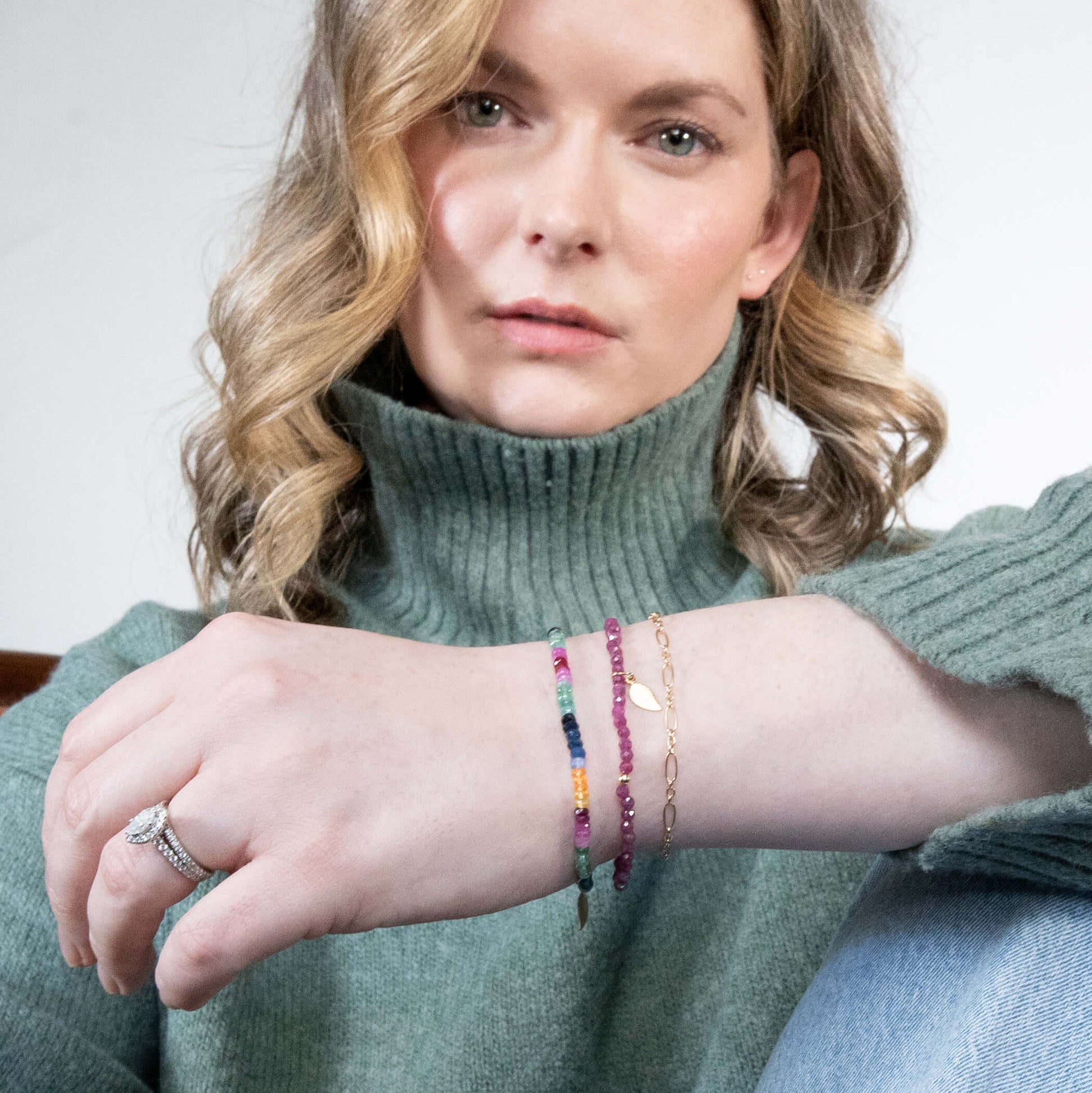 A woman wearing a sage green turtleneck sweater and light blue jeans, showcasing delicate beaded bracelets on her wrist. The bracelets include a multicolor rainbow bead strand, a pink beaded strand, and a thin gold chain with a small gold heart charm.