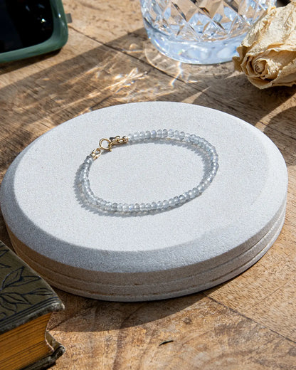 Grey with blue flash Labradorite faceted beaded bracelet with a small 14k gold-filled heart charm and spring ring clasp on a light coaster on wood table