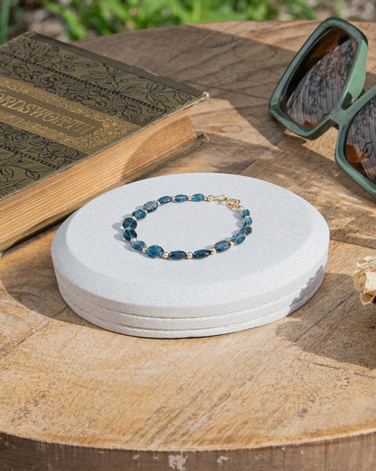 Dark blue London Topaz faceted oval-shaped beaded gemstone bracelet with 14k gold-filled accent beads and spring ring clasp on light coaster on wood table