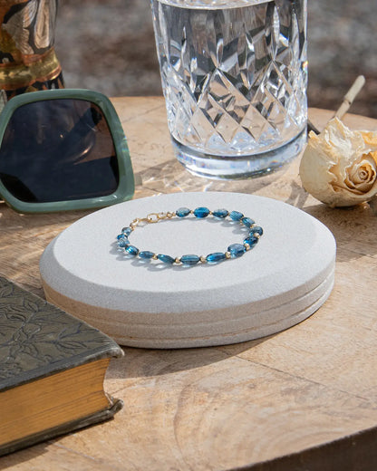 Dark blue London Topaz faceted oval-shaped beaded gemstone bracelet with 14k gold-filled accent beads and spring ring clasp on light coaster on wood table