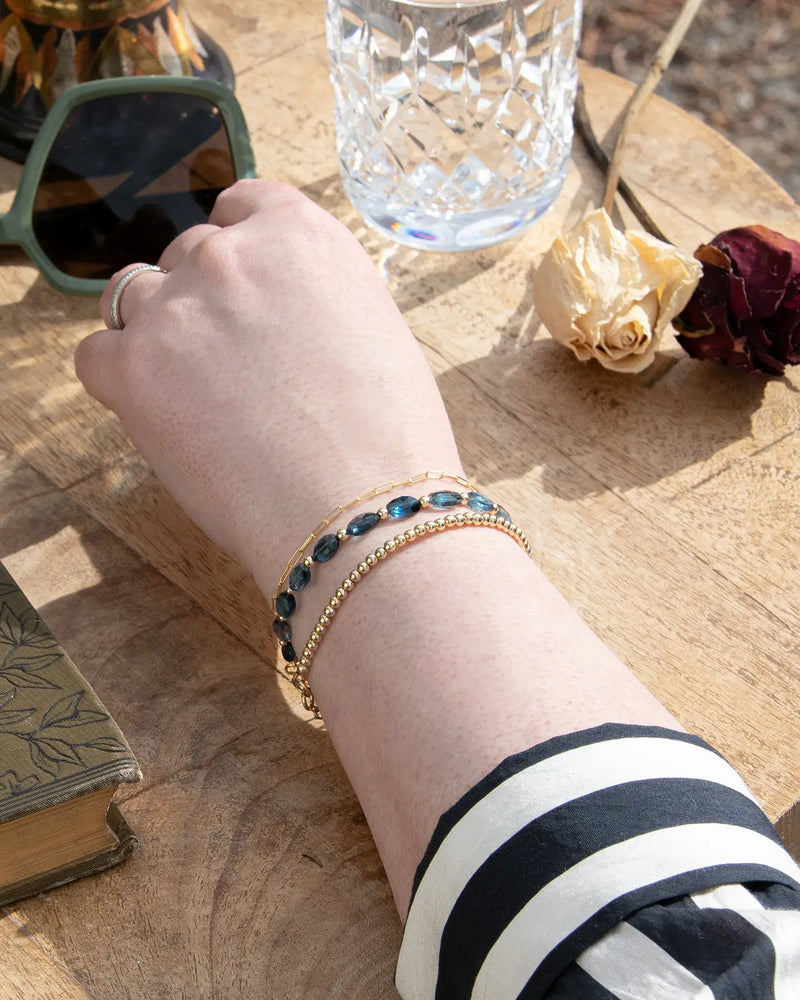 Dark blue London Topaz faceted oval-shaped beaded gemstone bracelet with 14k gold-filled accent beads stack on wrist with 14k gold-filled beaded bracelet and 14k gold-filled paperclip chain on wood table
