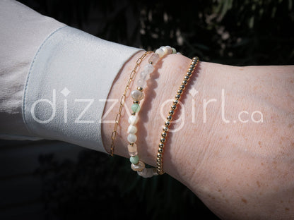A beaded bracelet with pearlescent white mother of pearl beads, oval-shaped freshwater pearl, opaque white quartz, and pale green jade separated by 14k gold-filled accent beads, stacked on a wrist with a 14k gold-filled beaded bracelet and 14k gold-filled paperclip chain