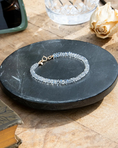 Clear white Moonstone faceted beaded bracelet with flashes of blue with small 14k gold-filled heart charm and spring ring clasp on black coaster on wood table