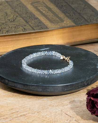 Clear white Moonstone faceted beaded bracelet with flashes of blue with small 14k gold-filled heart charm and spring ring clasp on black coaster on wood table