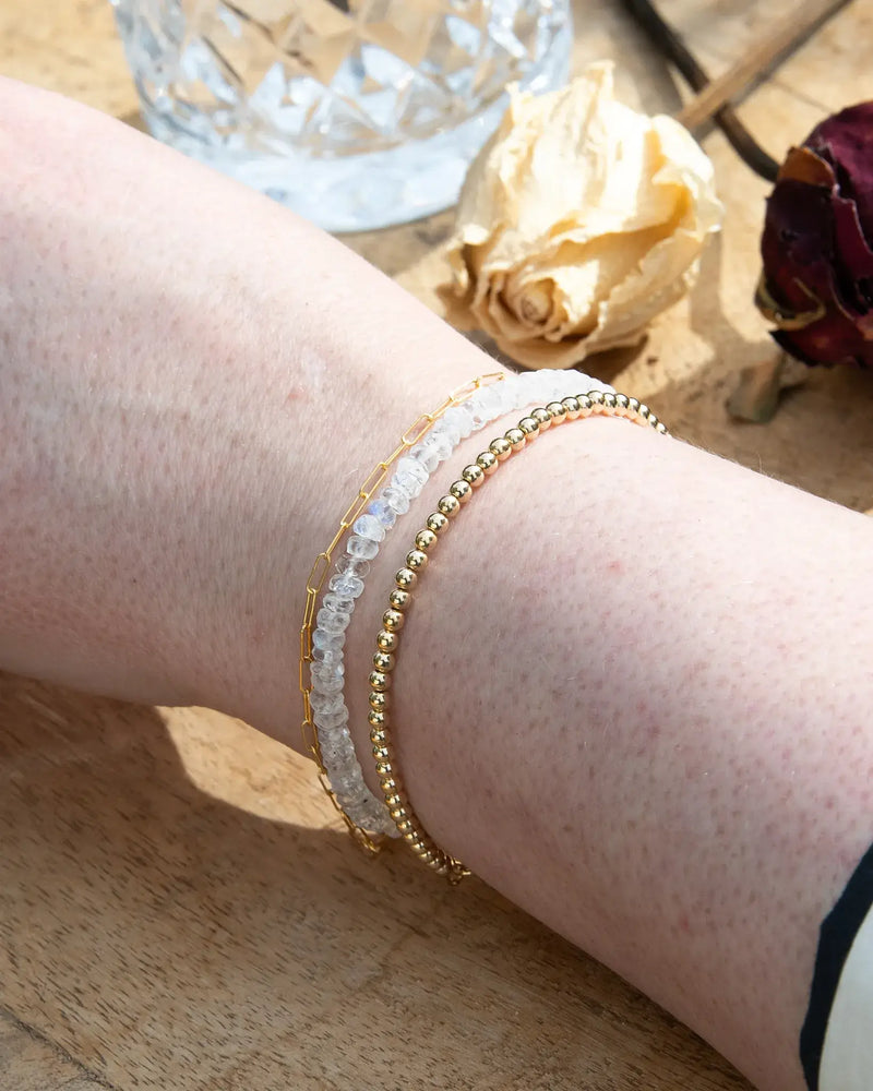Clear white Moonstone faceted beaded bracelet with flashes of blue stacked on wrist with with 14k gold-filled beaded bracelet and 14k gold-filled paperclip chain on wood table