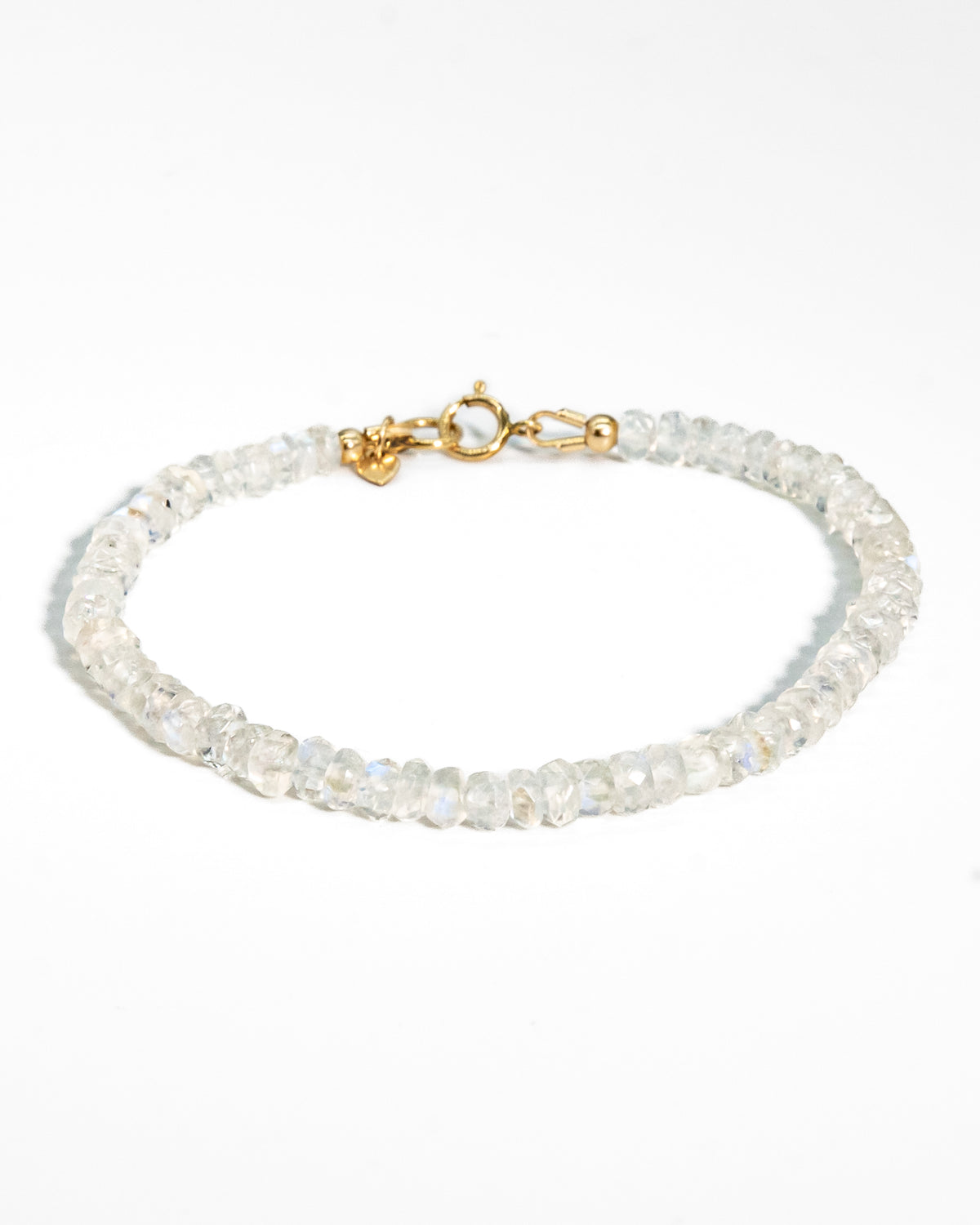 Clear white Moonstone faceted beaded bracelet with flashes of blue with small 14k gold-filled heart charm and spring ring clasp on black coaster on wood table
