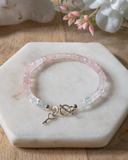 Pale pink, high clarity faceted morganite beaded bracelet with sterling silver key-shaped charm and toggle closure on a white  light coaster on wood table