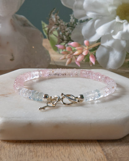 Pale pink, high clarity faceted morganite beaded bracelet with sterling silver key-shaped charm and toggle closure on a white  light coaster on wood table