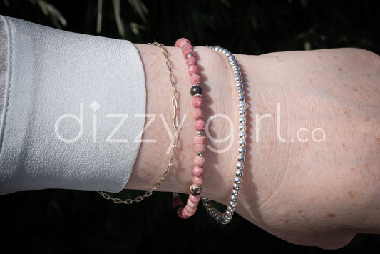 A Pink Rhodonite beaded bracelet with rosy-pink faceted round and smooth Heishi beads separated by black rhodonite round beads and sterling silver accents, stacked on a wrist with a sterling silver beaded bracelet and a sterling silver paperclip chain