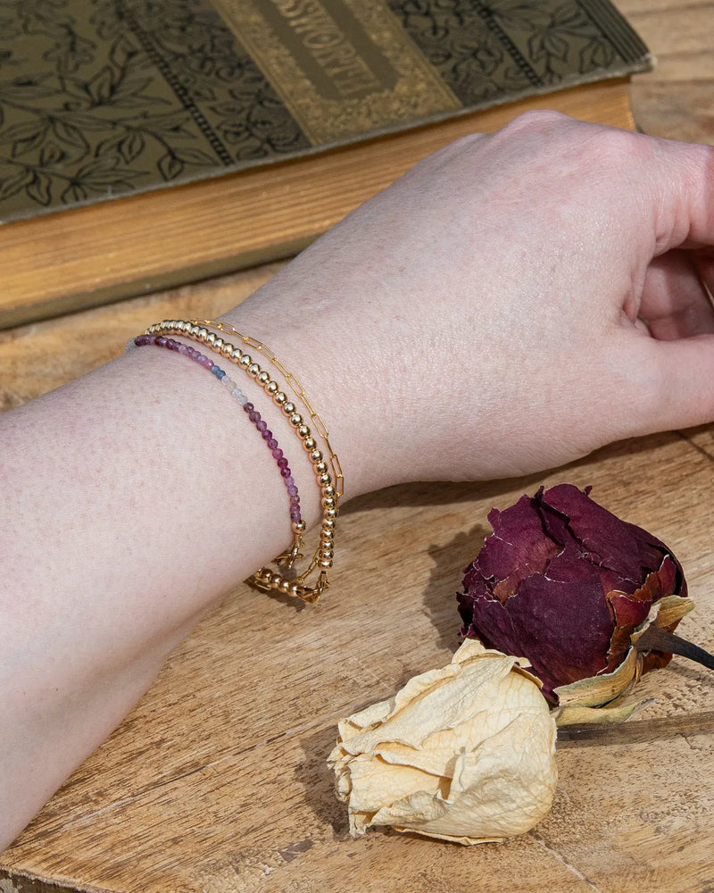 Dainty pink, blue, and white ruby sapphire beaded bracelet on wrist stacked with 14k gold-filled beaded bracelet and 14k gold-filled chain.